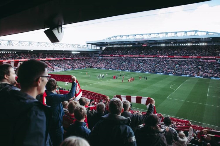 View of Anfield Stadium from the stalls filled with fans holding up their scarves and cheering in support of the players on the pitch